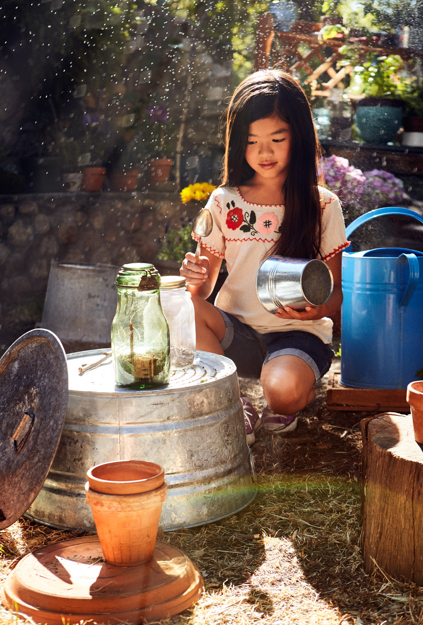 Girl in embroidered top playing in garden with glass jars and watering can, Amazon Kids lifestyle photography