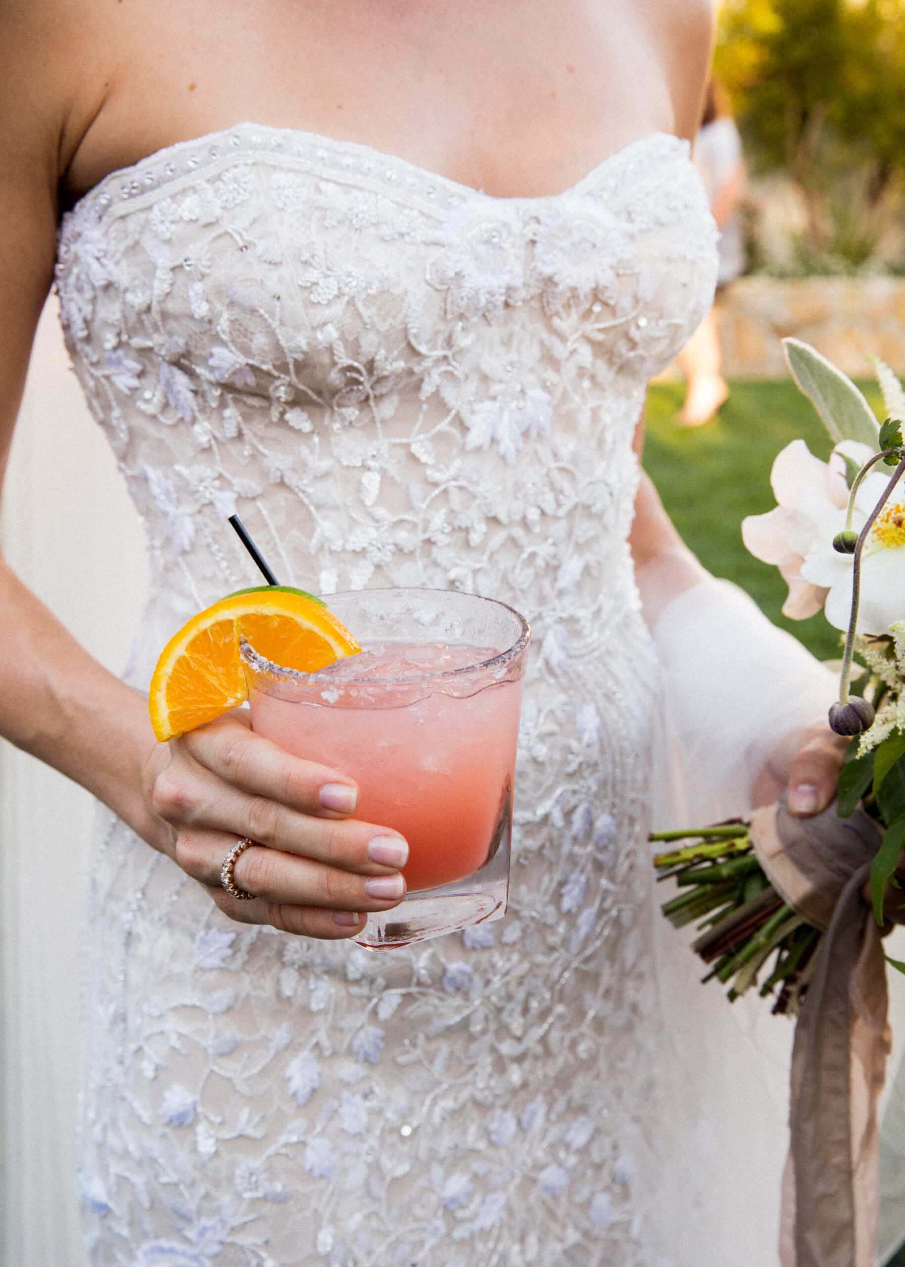 Bride in lace strapless gown holding citrus cocktail and bouquet, luxury wedding detail