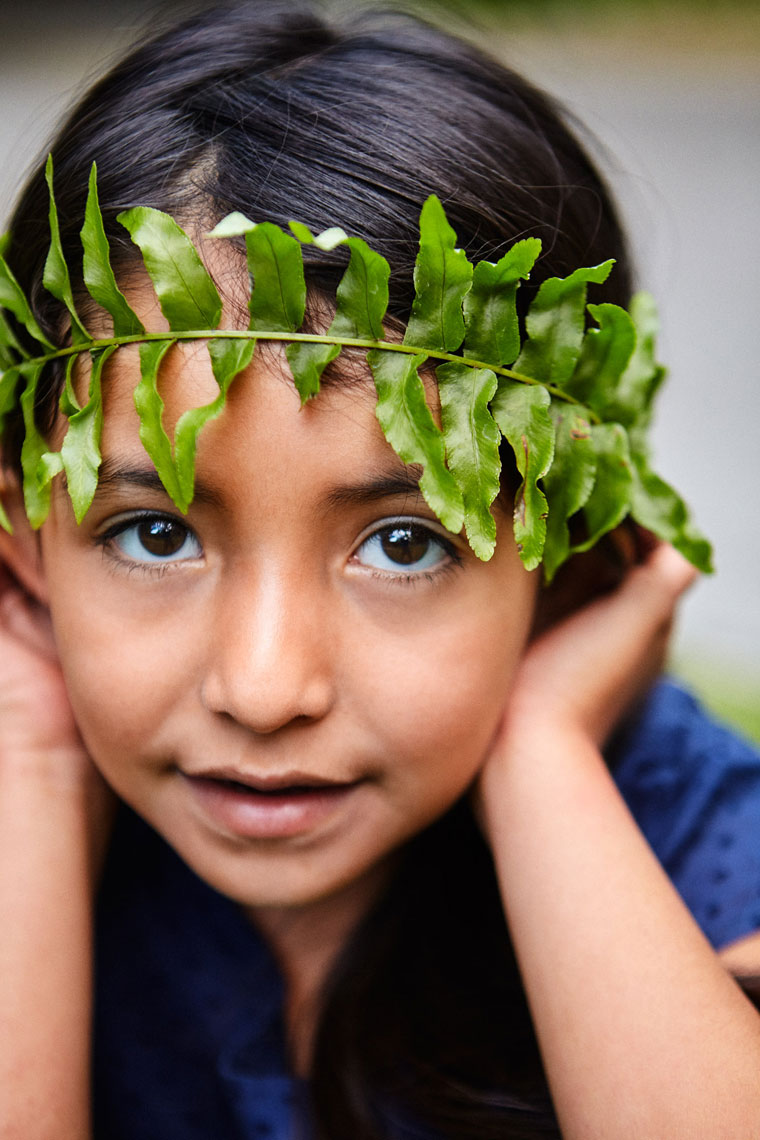 Child with fern vine crown holding face in hands, natural light editorial portrait photography
