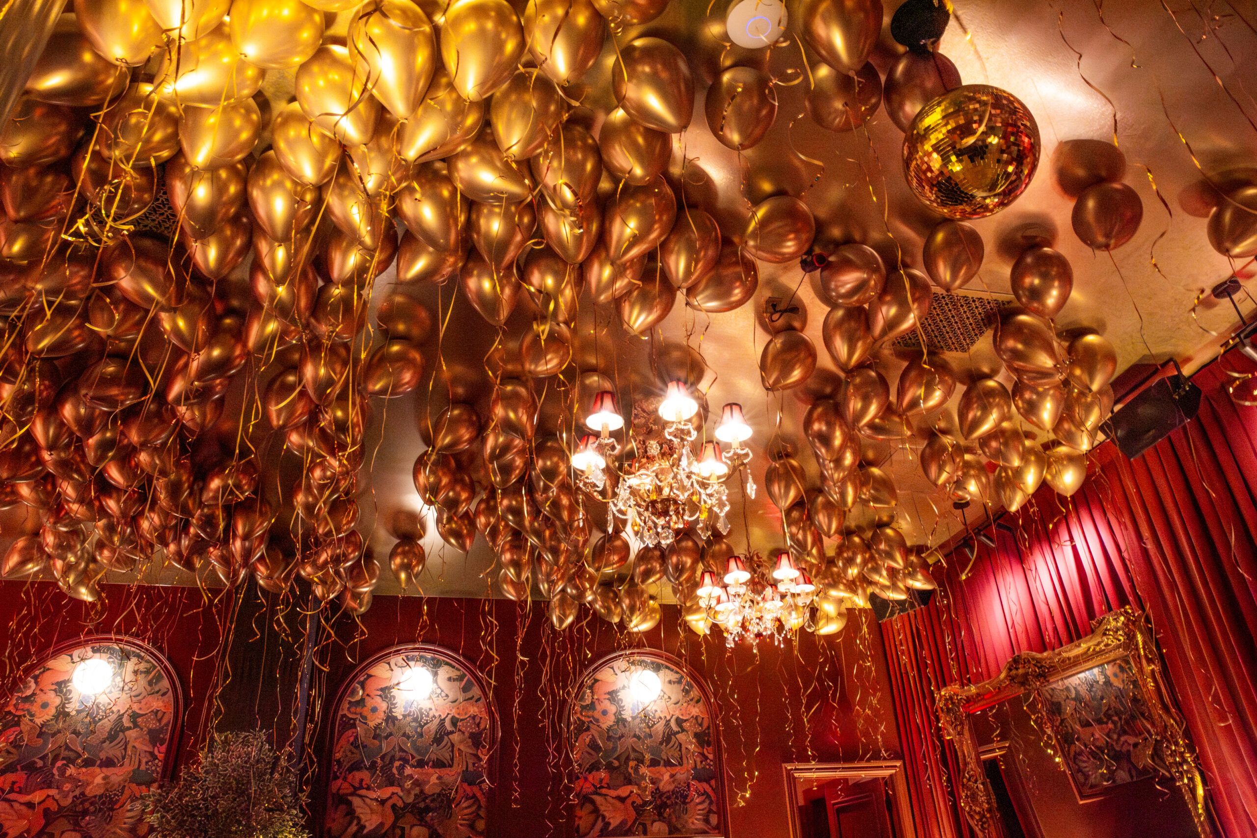 Gold chrome balloons covering ceiling with crystal chandeliers and red velvet walls, luxury event production photography