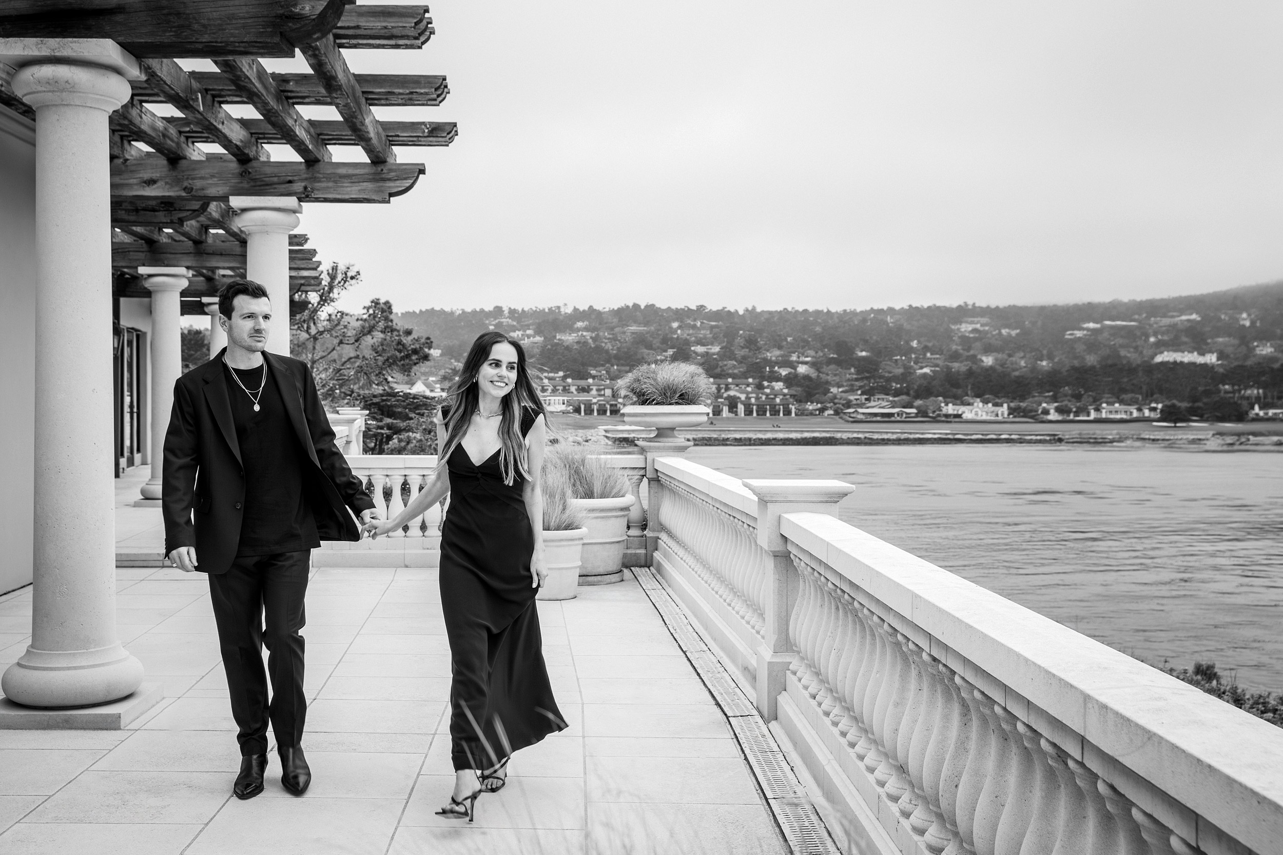 Couple in black walking hand in hand along stone terrace colonnade overlooking Pebble Beach bay, black and white luxury lifestyle editorial photography