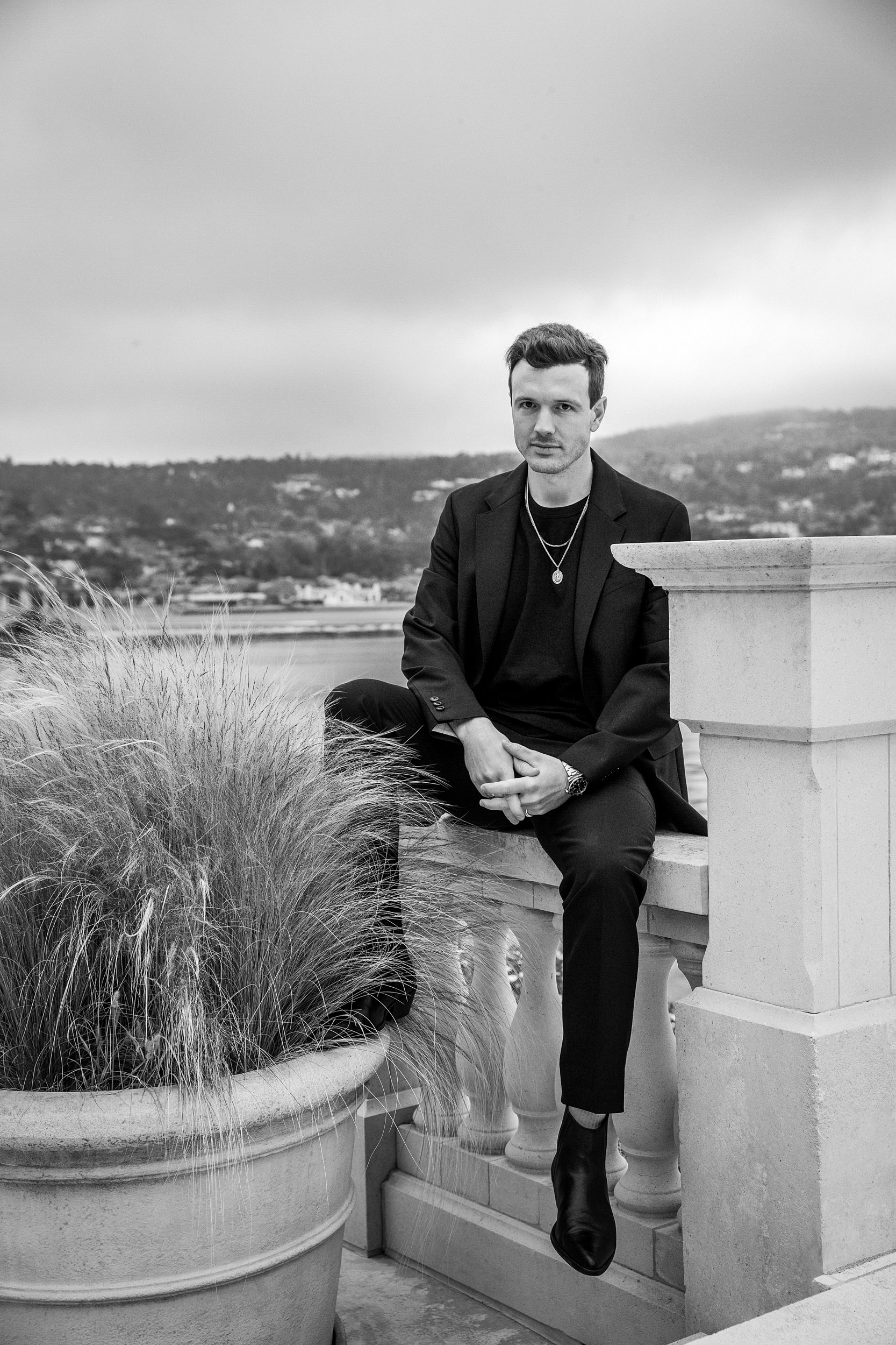 Man in black seated on stone balustrade overlooking Pebble Beach bay, black and white luxury lifestyle editorial photography