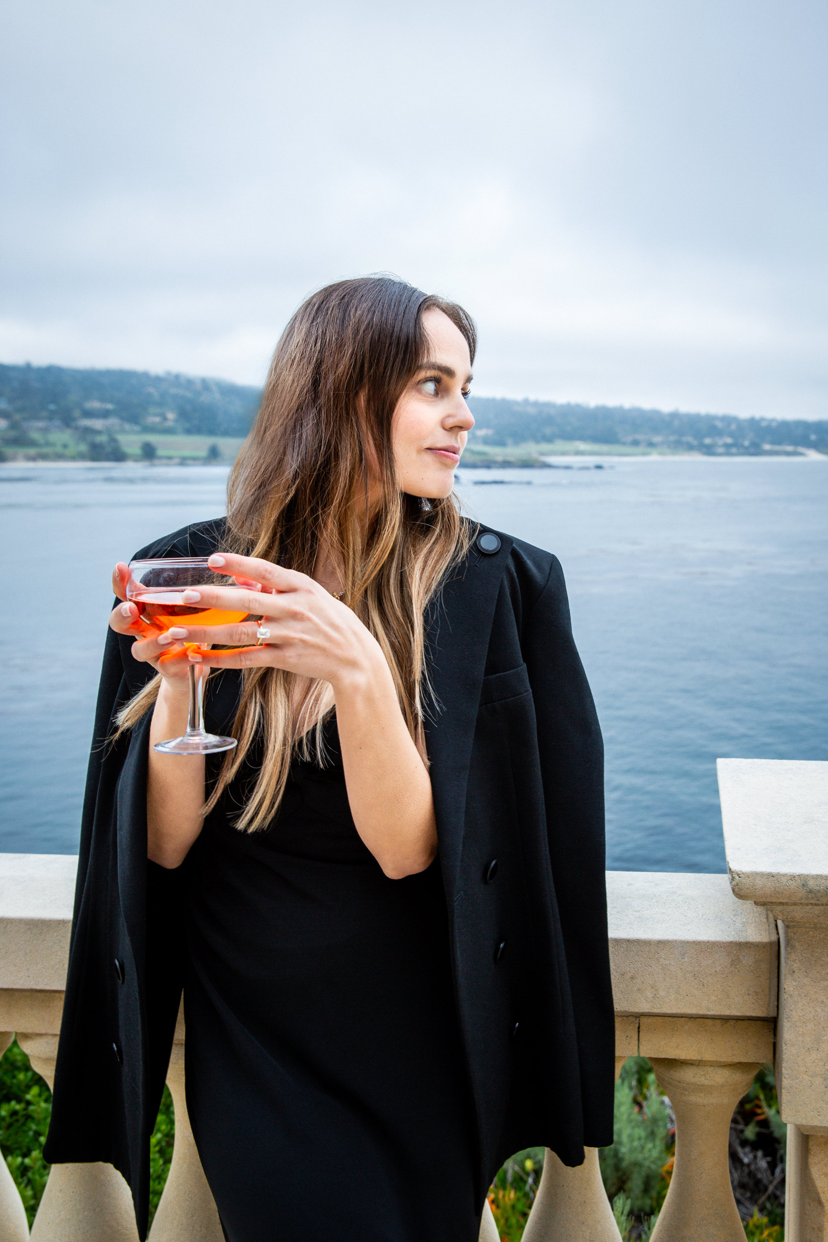 Woman in black blazer holding aperol coupe on stone terrace overlooking Pebble Beach bay, luxury lifestyle editorial photography