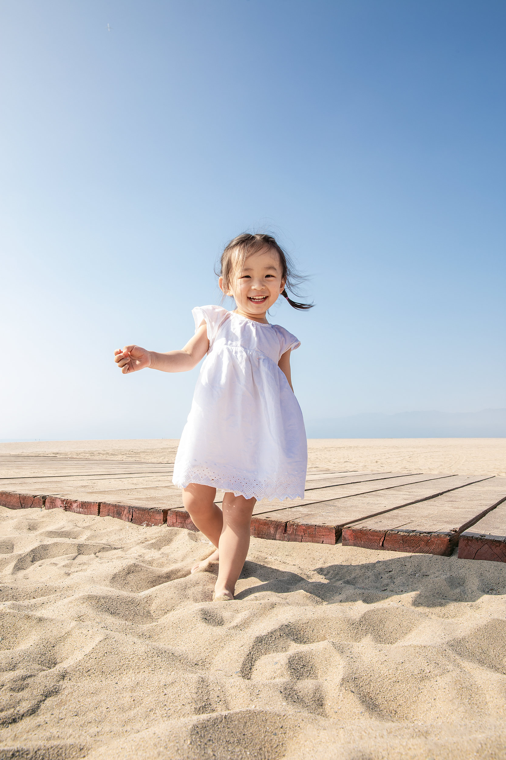 young girl white dress arms outstretched running beach blue sky joyful lifestyle editorial photography